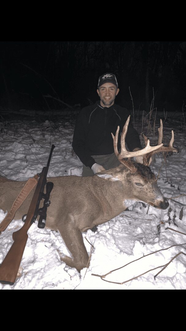 A man with a hunting rifle posing with a dead deer in the snow