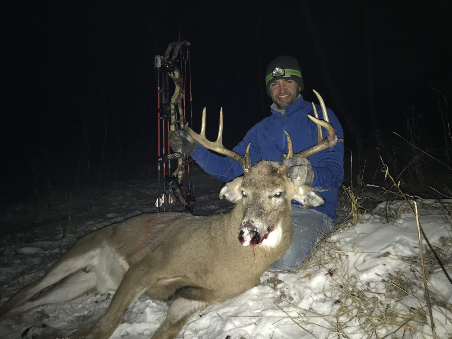 A man with a hunting bow posing with a dead deer in the snow