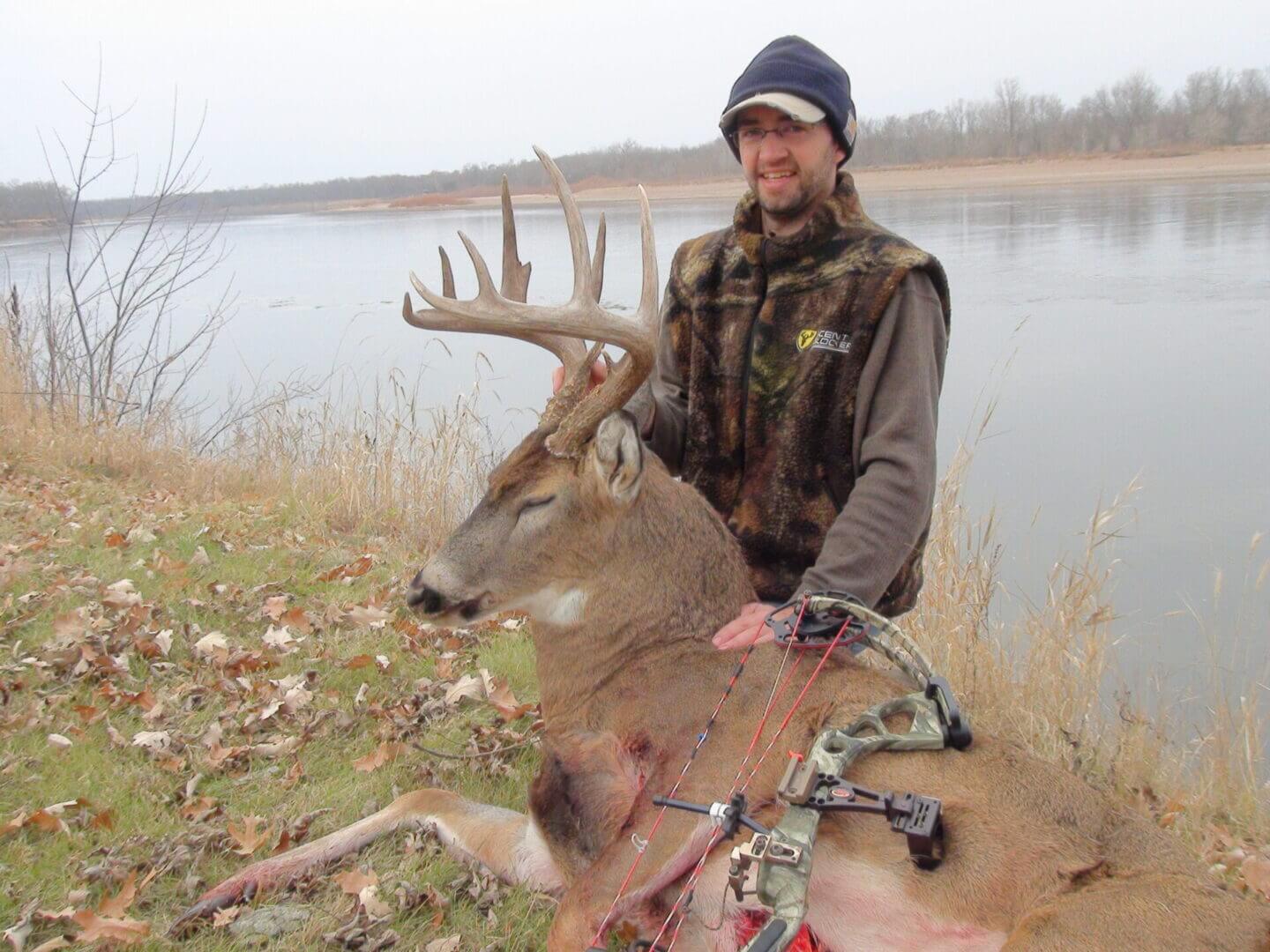 A man with a camouflage vest posing with a dead deer